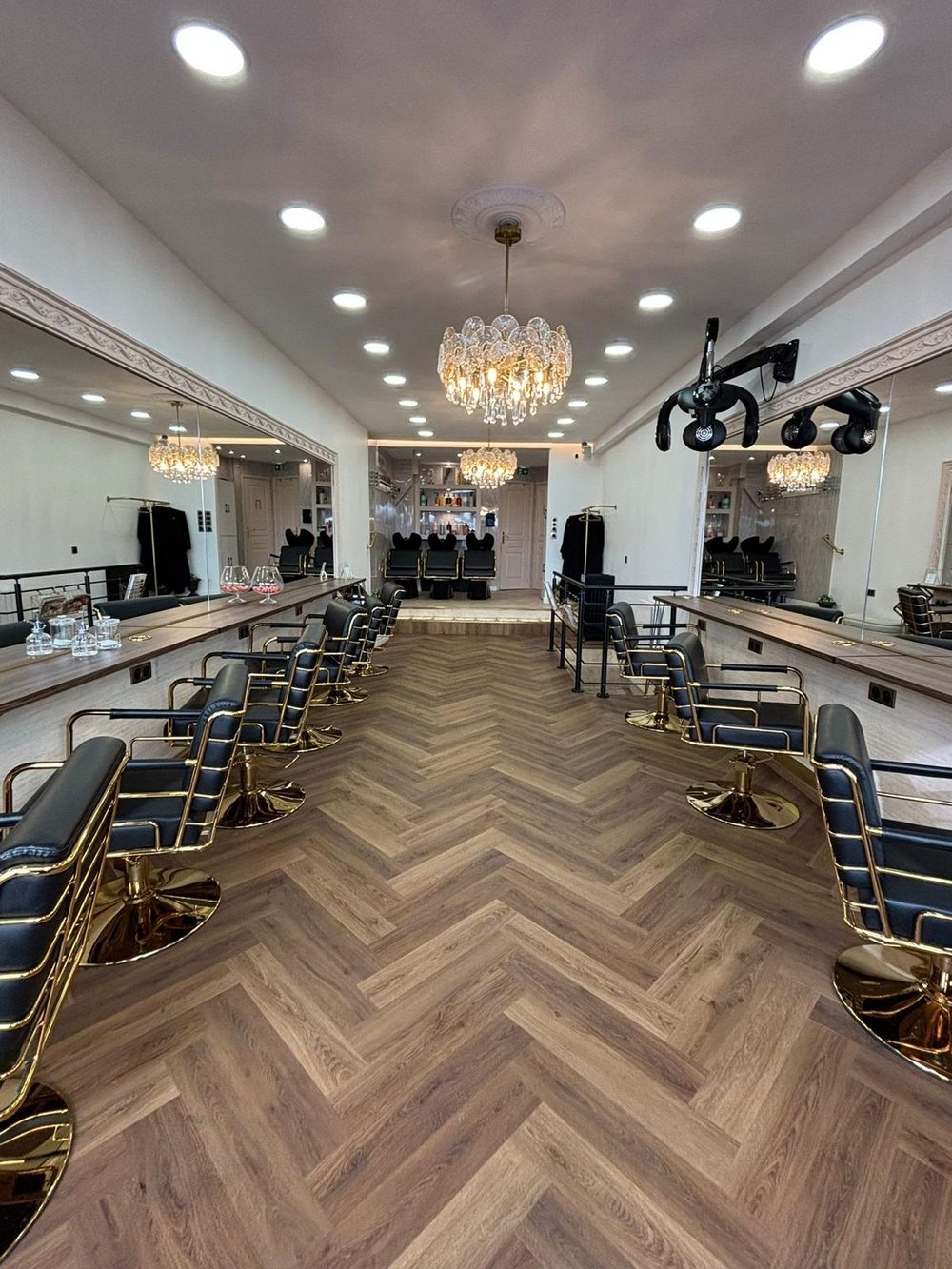 La salle de coiffure Rosana : postes en enfilade, fauteuils noir et or, lustre en cristal et parquet à chevrons.